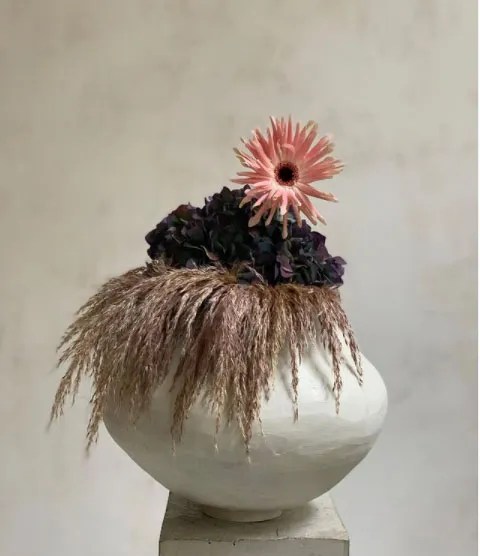 Paris based Castor flowers arrangement with single pink flower amongst dark blue and light purple hay in white vase Paris based Castor flowers arrangement with single pink flower amongst dark blue and light purple hay in white vase